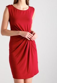 Röd ärmlös klänning med rund halsringning, tillverkad av ett slätt tyg, med en vriden framdesign och en figurnära siluett.