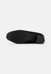Zapato de ante negro con suela texturizada y estrías horizontales, y un tacón redondeado y liso. Diseño simple adecuado para un uso versátil.