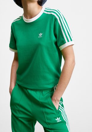 Persoon draagt een groen Adidas trainingspak met witte strepen op de mouwen en broekspijpen, handen in de zakken en een donkerblauwe hoed.