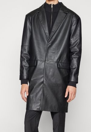 Homme portant un long manteau en cuir noir sur un sweat à zip noir et un pantalon sombre, se tenant devant un fond uni.