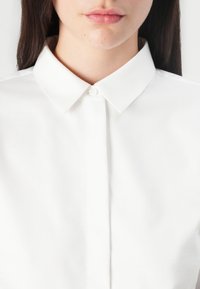 Chemise blanche à col classique avec un devant à boutons, présentant une texture lisse et un design intemporel, montrant un gros plan du col et de la patte de boutonnage.