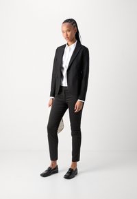 Černý přizpůsobený blazer přes bílou košili, v kombinaci s černými slim-fit kalhotami a mokasíny. Osoba drží světle zbarvenou kabelku.