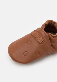 Pantof cu talpă moale din piele maro pentru bebeluși, cu model de urme de labe embosate, deschidere elastică și textură netedă. Soluție confortabilă pentru purtare.