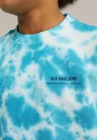 Šviesiai mėlynas ir baltas trumpomis rankovėmis marškinėliai su tinklelio raštu, kuriuose užrašyta "Blue Ridge Denim" ir "Nuotykiai prasideda ten, kur baigiasi miestas."