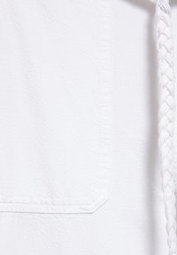 Gros plan d’un tissu blanc avec une poche rectangulaire cousue et un cordon blanc tressé le long du bord droit.