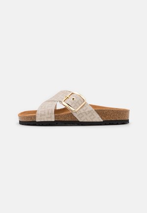Beige slidesandale med krydsede stofstropper, en guldbøjle, korkfodbed og sort tekstureret sål, vist fra siden på hvid baggrund.