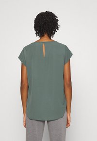 Zielona bluzka z rękawami typu cap, detal w kształcie klucza na plecach, zaokrąglony dół, wykonana z fakturowanej tkaniny, zestawiona z szarymi spodniami.