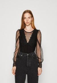 Body nero con maniche a pois trasparenti e accenti in pizzo, caratterizzato da un torso aderente e un colletto alto. Abbinato a jeans neri a vita alta.