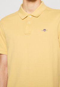 Polotröja i pastellgult, kortärmad, med en tre-knappars knäppning. Har en liten broderad logotyp på vänster bröst. Mjuk bomullstextur.