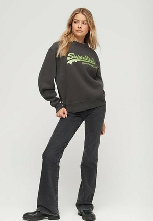 Felpa nera con logo "SuperDry" verde, abbinata a jeans neri a zampa d'elefante. Modello indossa stivali neri chunky, in piedi su uno sfondo neutro.