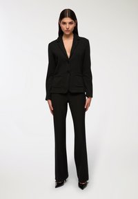 Blazer nero su misura con rever a punta, due tasche frontali e chiusura a un bottone. Abbinato a pantaloni fitted coordinati e tacchi a punta.