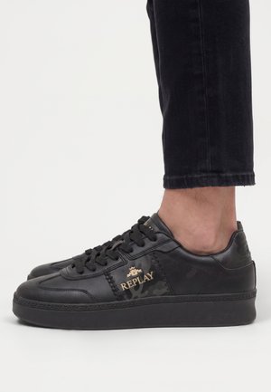 Persona che indossa sneaker nere del marchio Replay con logo dorato, abbinate a pantaloni neri corti su sfondo bianco semplice.