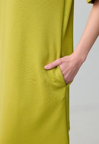 Abito in tessuto verde e morbido con una vestibilità rilassata, dotato di una tasca laterale, con una mano delicatamente appoggiata all'interno. Texture liscia, design semplice.