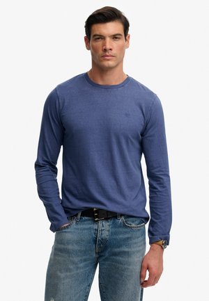 Hombre con camisa de manga larga azul, vaqueros azules desteñidos, cinturón negro y reloj de pulsera, de pie con una mano en el bolsillo.