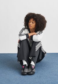 Ung kvinne med krøllete hår sitter med korslagte ben på blått teppe, iført svart-hvitt Adidas treningsdress og svarte joggesko med rosa logo.