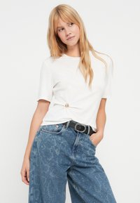 T-shirt blanc à manches courtes avec un détail de nœud sur le côté gauche, associé à un jean large bleu à motifs et une ceinture noire avec des accents argentés.