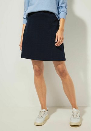 Woman wearing a black knee-length skirt, light blue long-sleeve top, and white sneakers, standing against a plain background.