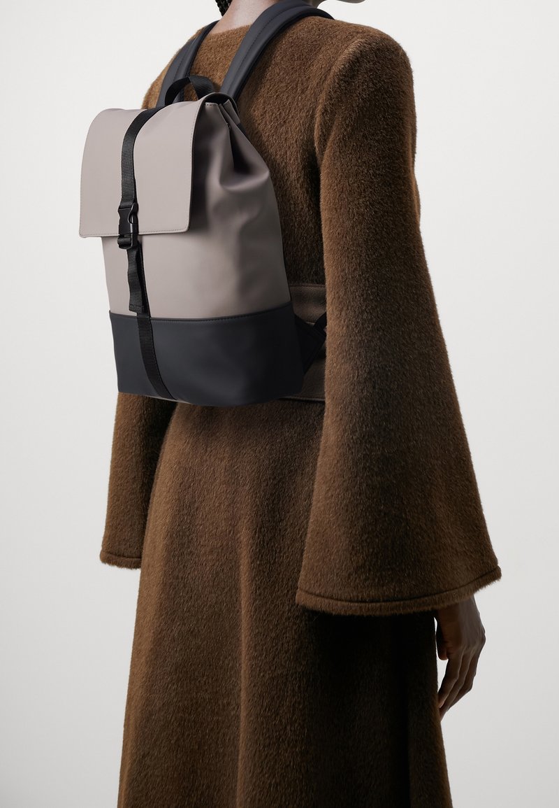 Un sac à dos bicolore gris et noir avec un rabat, des bretelles réglables et une texture mate, porté par-dessus un manteau marron.