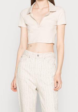 Vrouw die een beige cropped top met korte mouwen en kraag draagt, en een crème broek met hoge taille en een subtiel verticaal patroon.