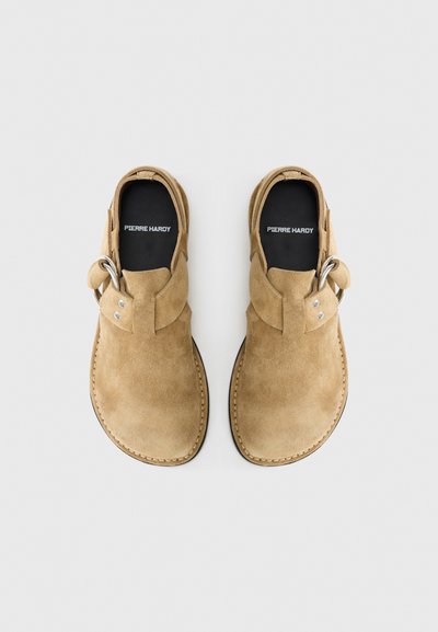 Καφέ παπούτσια από σουέντ με στρογγυλεμένη μύτη και ανοιχτές πλευρές. Διαθέτουν ασημένια αγκράφα και αντίθετη ραφή κατά μήκος της σόλας.