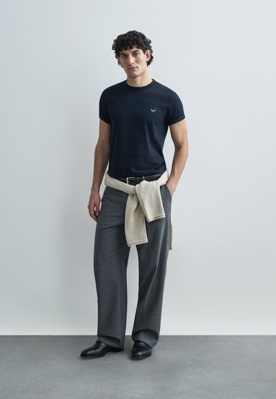 Tamsiai mėlynas aptemptas marškinėlius su logotipu, šviesiai smėlio spalvos megztinis, rišamas ties juosmeniu, pilkos platėjančios kelnės ir juodi odiniai batai. Lygios audinio tekstūros.