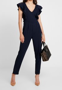 Námořnický modrý overal s volánovými rukávy, výstřihem do V, zúženými nohavicemi a záhyby. Nesený s černou a hnědou kabelkou s hadím vzorem.