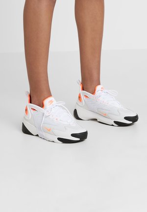 White and orange Nike sneakers with black soles worn on feet against plain light gray background.