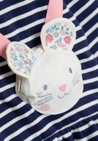 Balta audinio kišenė triušio formos su gėlių raštuotomis ausimis, rožinėmis juostelėmis ir mėlyna bei rožinė siuvinėjimu ant mėlynai baltų dryžuotų fono.