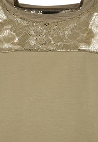 Haut beige avec encolure côtelée et empiècement en dentelle florale sur le haut de la poitrine et les épaules.