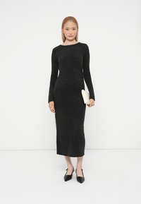 Črna pletena obleka dolgih rokavov, prilegajočega kroja, z okroglim izrezom in dolžino do tal. Ujemajoča se z elegantnimi črnimi petkami in belim clutchom.