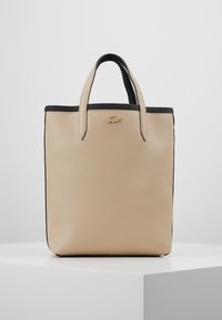 Sac tote en cuir beige avec des finitions noires, deux anses et une surface texturée. Logo Lacoste embossé à l'avant. Forme rectangulaire.