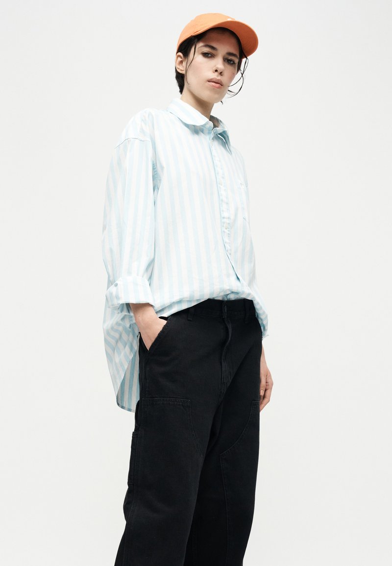 Jeune personne portant une casquette orange, une chemise surdimensionnée à rayures bleu clair et blanc, et un pantalon noir ample, les mains dans les poches, debout devant un fond blanc.