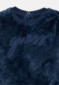 Tamno plavi plišani dukserica s mekanom teksturom, s izvezenim "guess" logotipom u odgovarajućoj boji. Okrugli izrez i detalj s petljom na leđima.