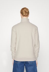 Persona con capelli biondi raccolti in uno chignon, che indossa un maglione beige a collo alto e jeans blu, di spalle contro uno sfondo bianco semplice.