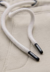 Gros plan sur des cordons de capuche tressés blancs avec des embouts en plastique noir portant l'inscription "DIMBRE" sur un tissu beige.