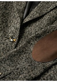 Tekstureret grå og sort jakke med en glat brun semsket læderalbuelap, to knapper og et klassisk kravedesign.