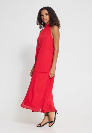 Vrouw met krullend haar, gekleed in een rode mouwloze maxi-jurk en zwarte sandalen met lage hak, staand tegen een witte achtergrond.
