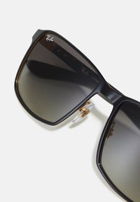 Ochelari de soare pătrați negri cu lentile gri degradé, accente aurii și un tampon de nas transparent. Prezentă branding-ul Ray-Ban pe ramă.