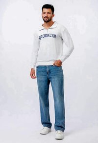 Sweatshirt cinza claro com o texto "BROOKLYN", colarinho com fecho, combinado com jeans azuis e ténis brancos. O tecido parece suave e descontraído.