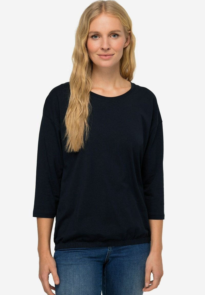 Černý, volný příjemný top s dlouhým rukávem a zaobleným výstřihem, se staženým lemem, vyrobený z hladkého materiálu. Doplňuje ho modré džíny.