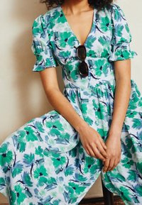 Kvinna klädd i en grön och blå blommig klänning med puffärmar, knappar fram och solglasögon hängande på halslinningen.