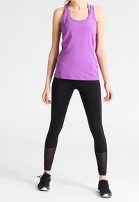 Lila atletisk tank top med breda axelremmar, parat med svarta leggings med texturerade meshdetaljer vid ärmsluten. Svarta sneakers fulländar outfiten.