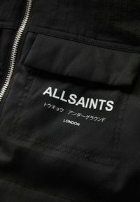Melna auduma jaka ar rāvējslēdzēju, kabatu ar uzšuvēm un tekstu: "ALLSAINTS" baltā krāsā, kā arī papildu tekstu japāņu valodā un "LONDON".