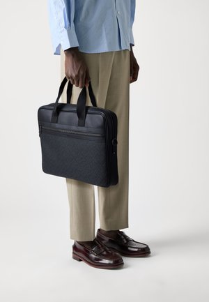 Black laptop bag with a textured front, dual handles, and a zippered compartment. Worn with beige trousers and brown shoes.