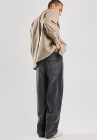 Camicia oversize beige con polsini a bottone, abbinata a jeans neri ampi con tasche posteriori e un aspetto sbiadito. Il modello sta in piedi con le mani nelle tasche.