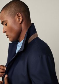 Marinejacke mit hohem Kragen, versehen mit einem kontrastierenden, beige Akzent an den Schultern, Reißverschluss und einer glatten, robusten Stofftextur.