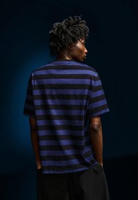 Tričko s krátkým rukávem a kulatým výstřihem se vodorovnými černými a fialovými pruhy. Vyrobeno z bavlny, má volný střih a měkkou texturu.