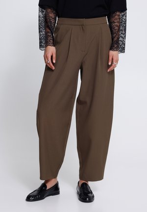Persona che indossa pantaloni marroni a gamba larga, mocassini neri e un top nero con maniche lunghe trasparenti in pizzo, in piedi contro uno sfondo semplice.