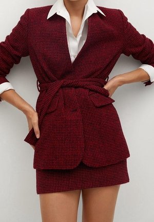 Vrouw draagt een rood-zwart geruit colbert met ceintuur in de taille en een bijpassend minirokje, gecombineerd met een wit overhemd met kraag eronder.