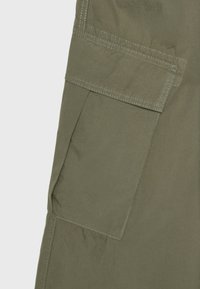 Gros plan d'une poche cargo en tissu vert olive avec un rabat rectangulaire et des coutures visibles.